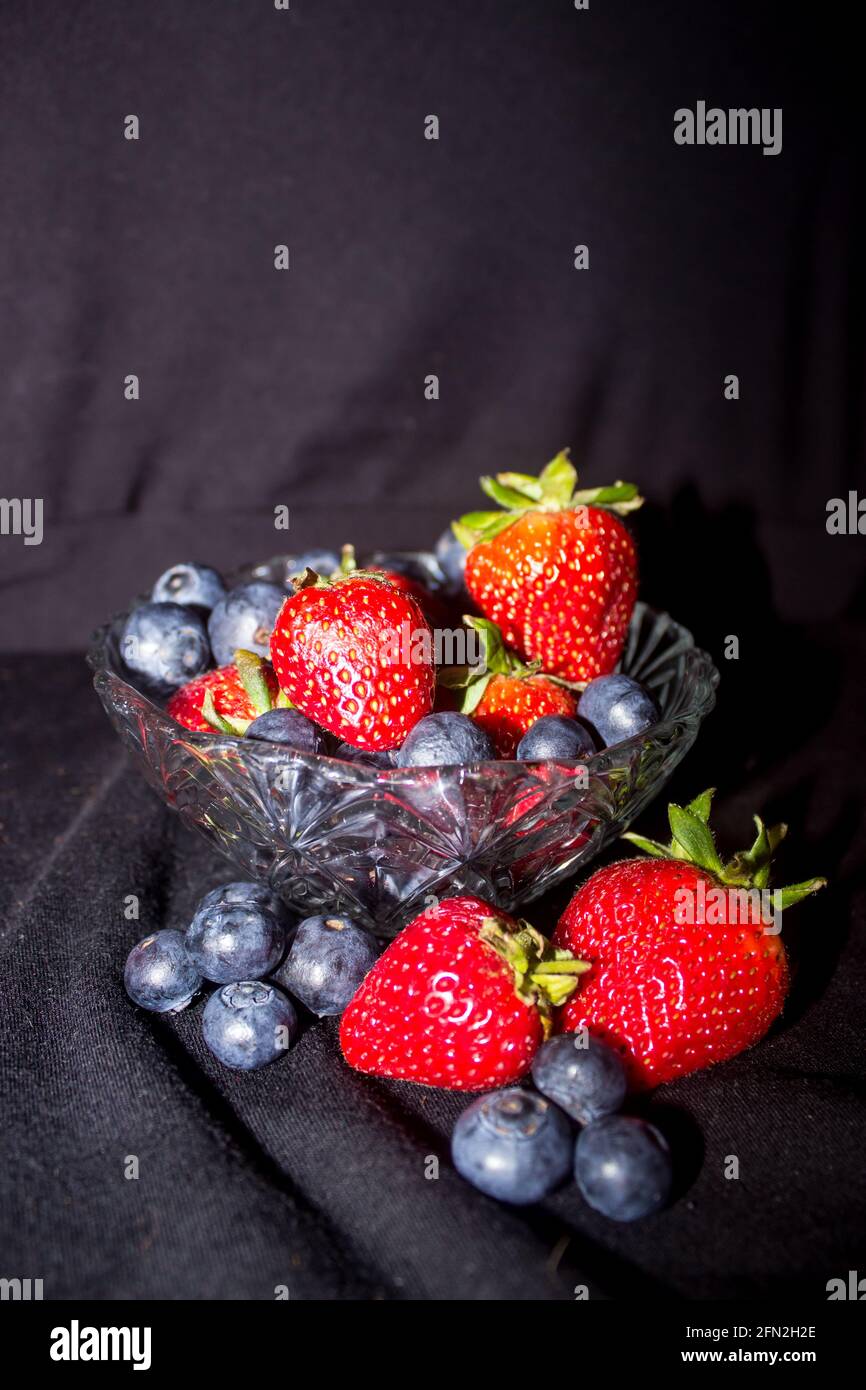 A small glass bowl filled and surrounded with a mixture of blueberries and strawberries, against a black background Stock Photo