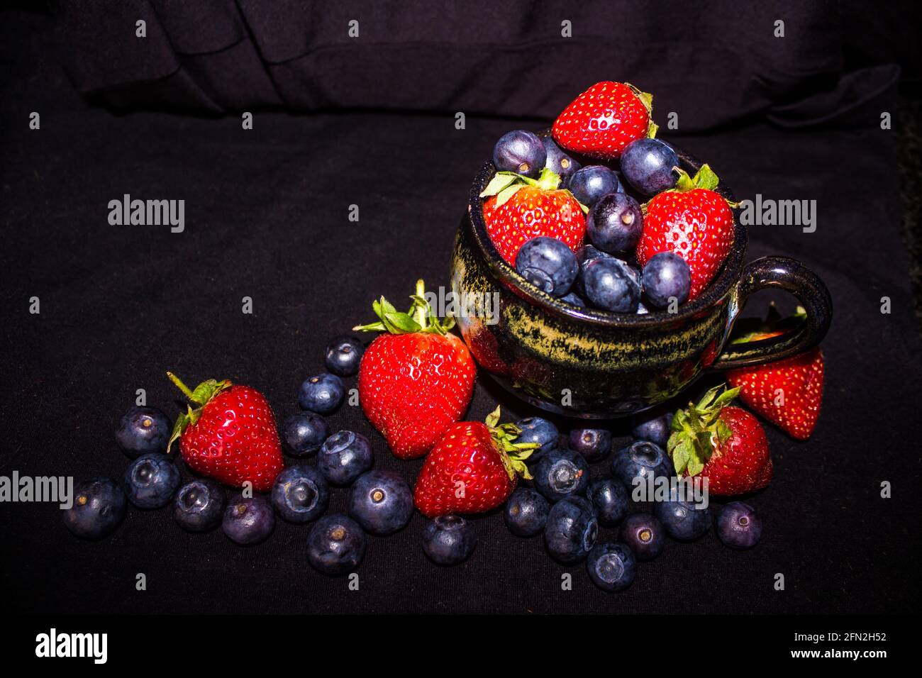 A still-life consisting of a black teacup filled to overflowing with strawberries and blueberries, with more surrounding it Stock Photo