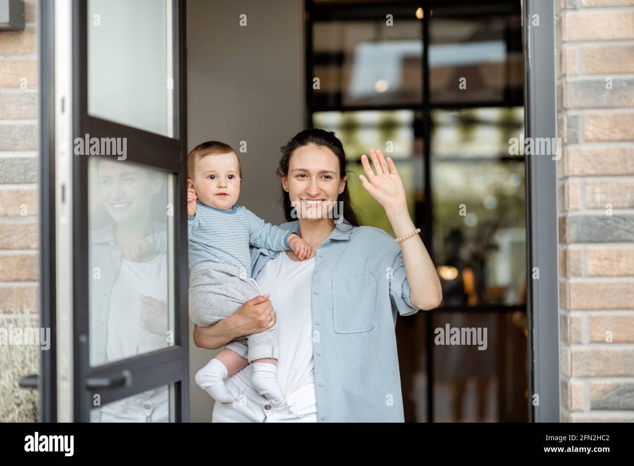 Housewife with newborn baby staying in front of entrance and waving