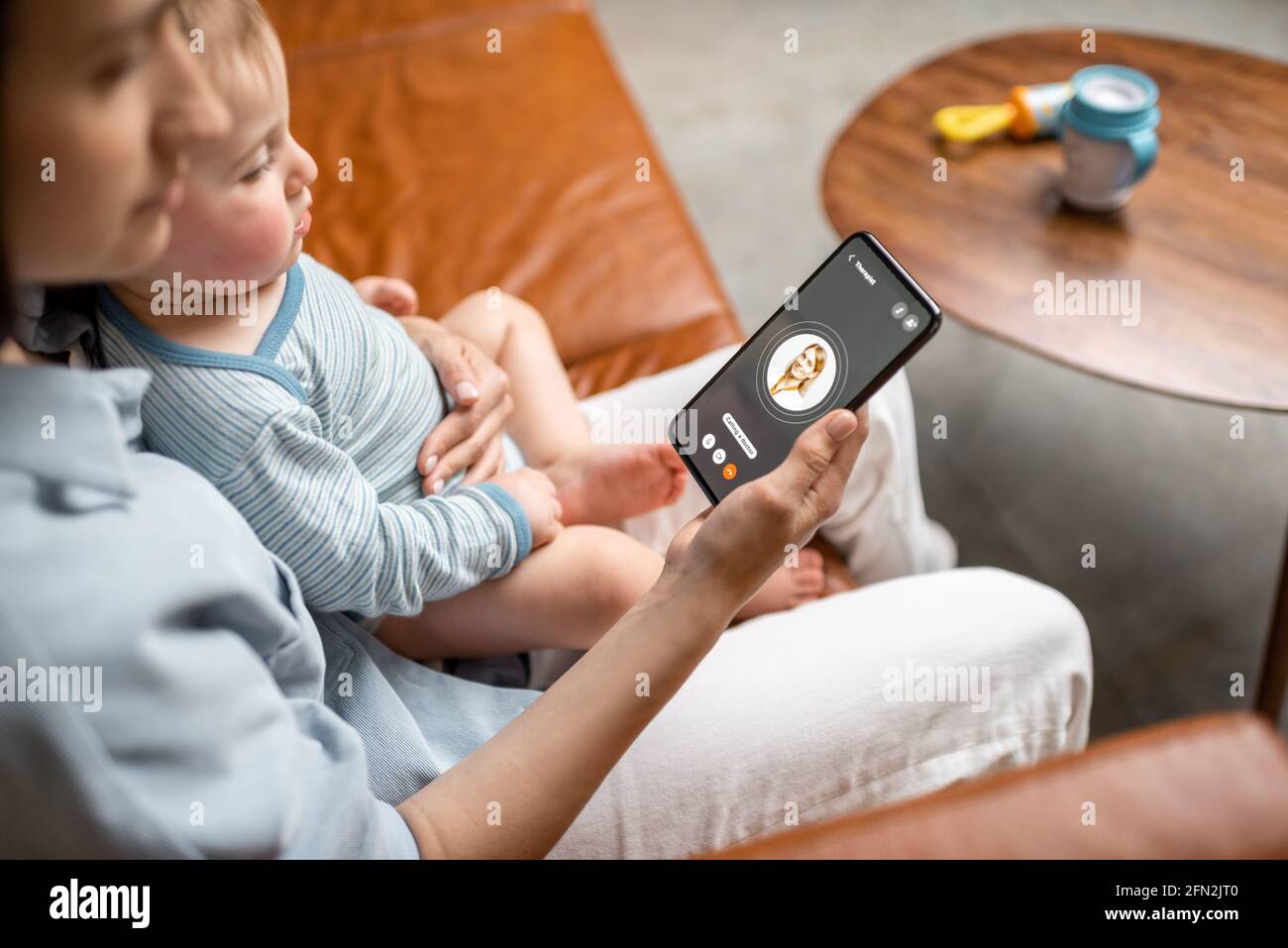 Young mother with newborn kid making video call to a doctor using phone at home. Concept of telemedicine and support parents counseling online.  Stock Photo