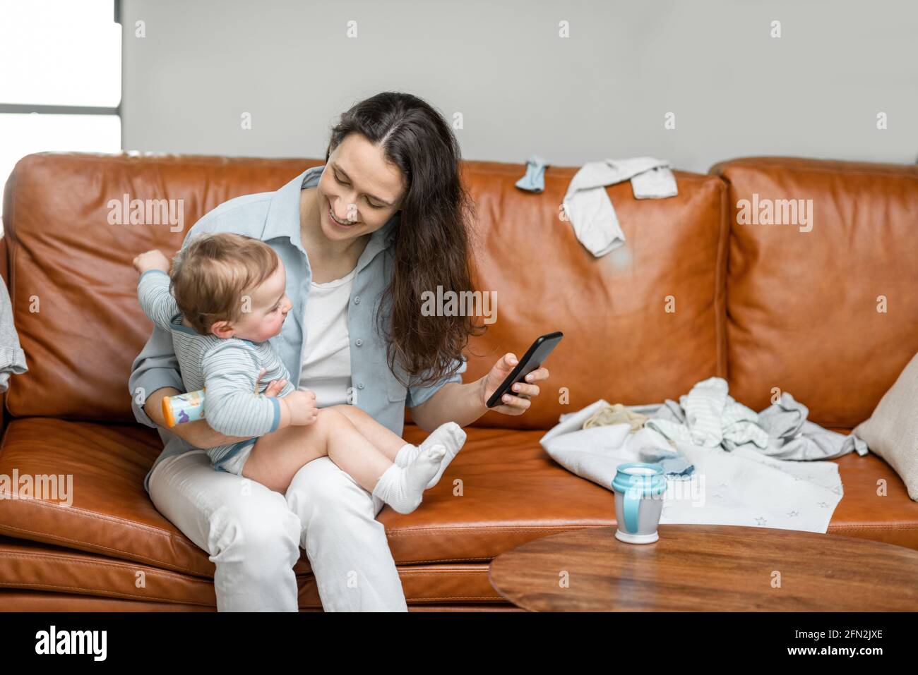 Young mom sitting with newborn son on sofa and take care or playing with a child. Raising baby, babysitter and motherhood. Entertain the child with the phone. Stock Photo