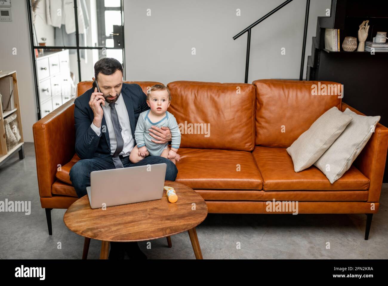 Young dad working on a laptop and speaking on phone at home while taking care of his baby son. Multitasking and babysitter. Stock Photo