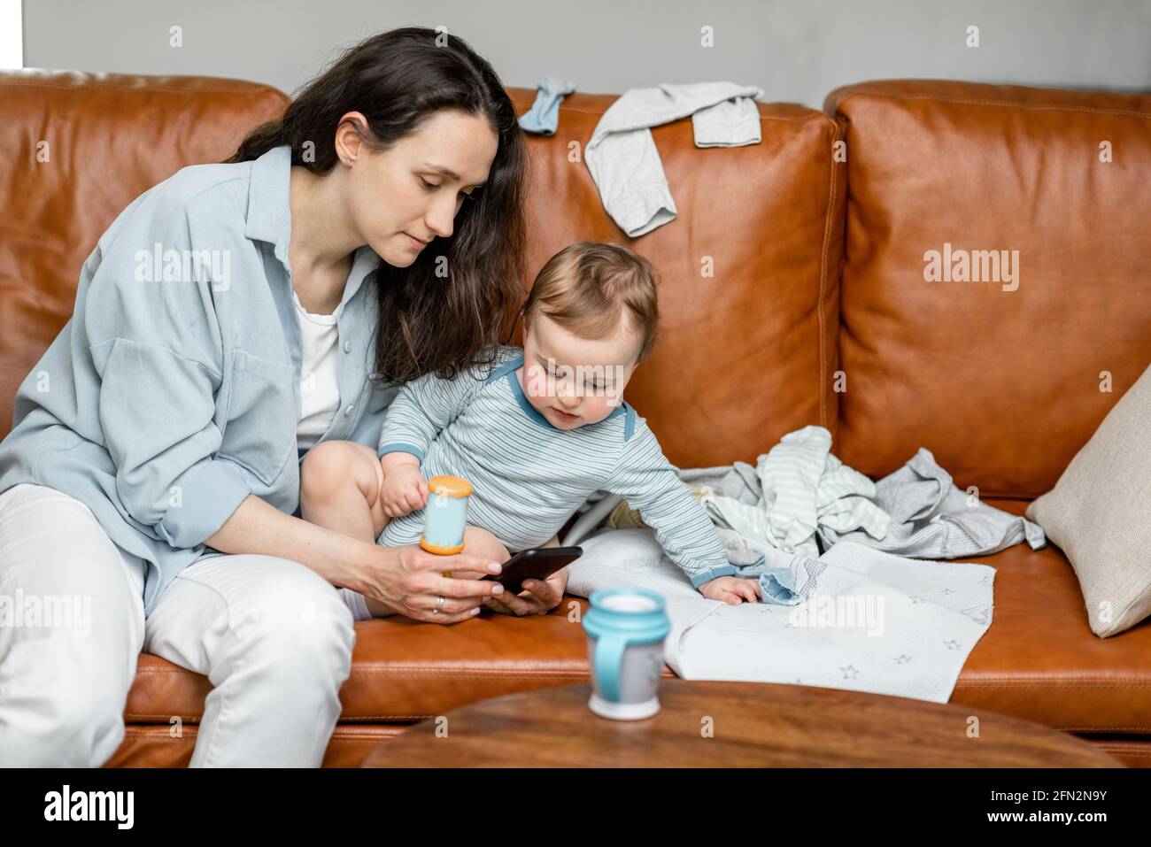Young mom sitting with newborn son on sofa and take care or playing with a child. Raising baby, babysitter and motherhood. Entertain the child with the phone. Stock Photo