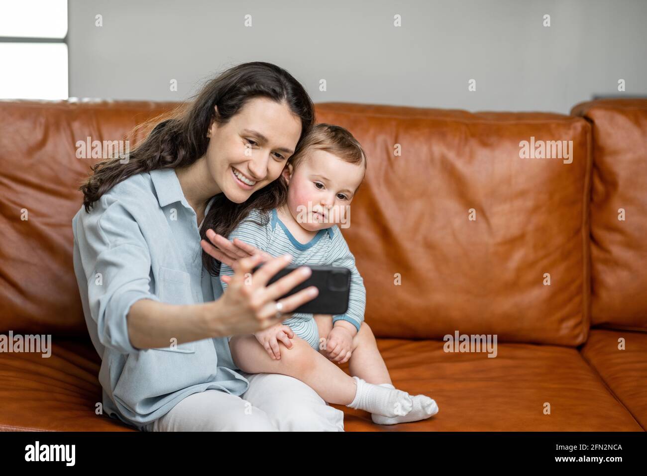 Young mom sitting with newborn son on sofa and making a video call or making a self with baby. Entertain the child with the phone. Stock Photo
