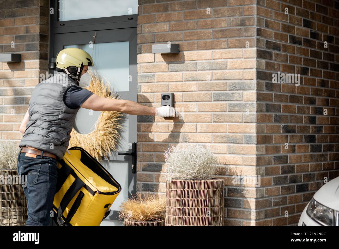 Courier in green helmet wearing mask and gloves holds a parcel box and  staying with yellow bag near front door of house. Ring the door bell and  waiting for customer to come