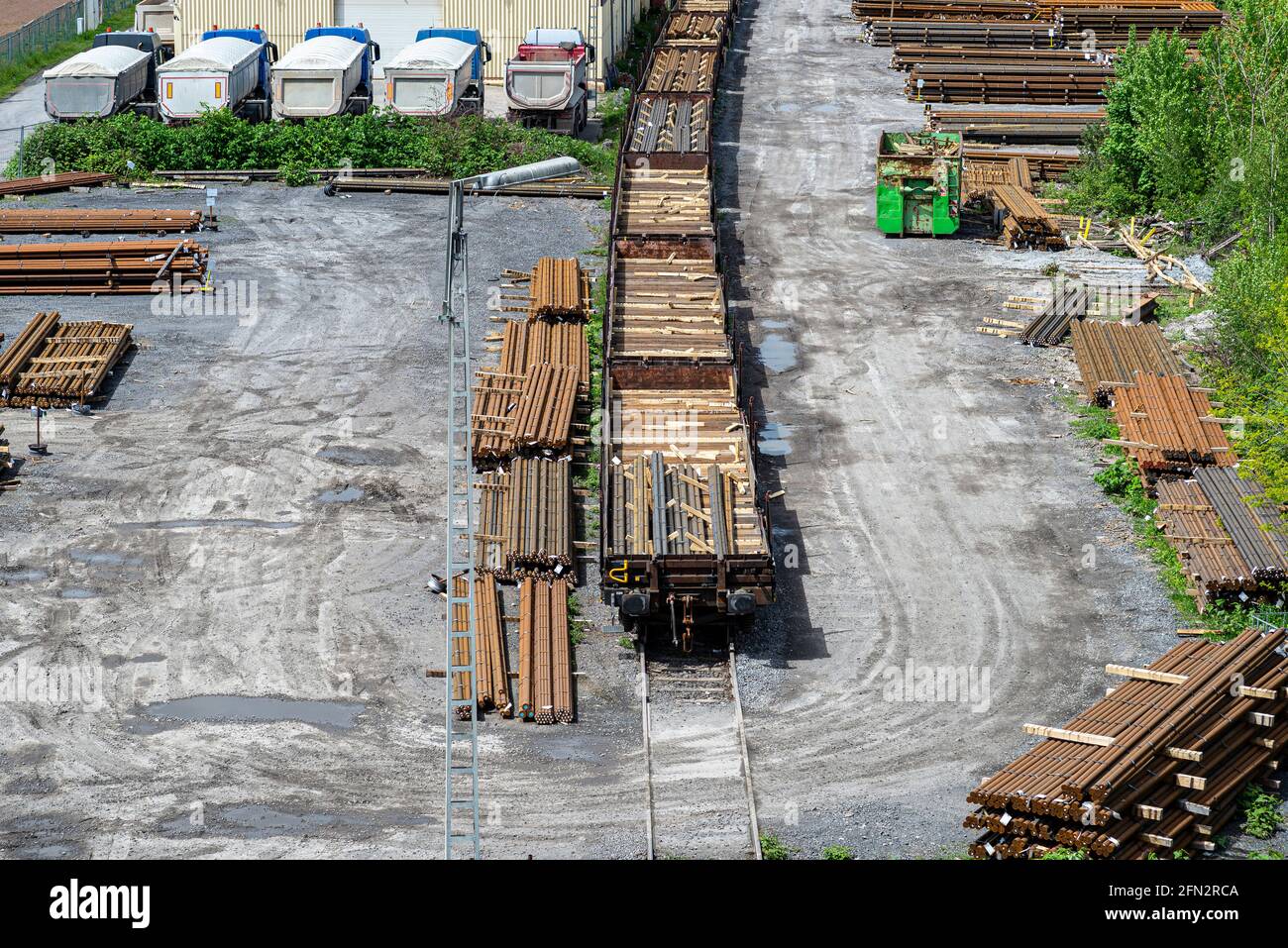 Long and thick, rust-covered steel bars stacked outside on a gravel square, visible railroad tracks and wagons, viewed from above. Stock Photo