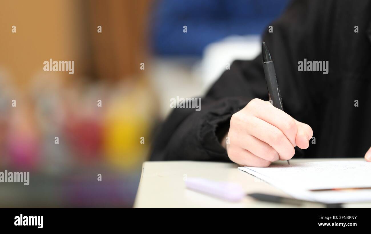 A close up isolated students hand holding a pen writing during class, exam or lesson time at school. Bright colors and second student blurred in the b Stock Photo