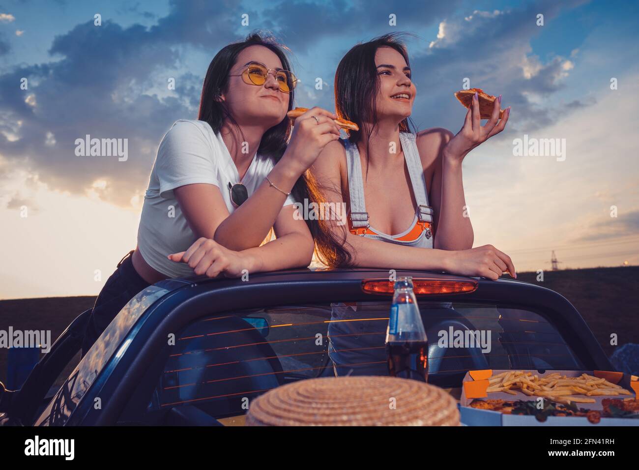Two women eating pizza, smiling, posing in yellow car cabrio with french fries, hat and soda in glass bottle on trunk. Fast food. Close up, mock up Stock Photo
