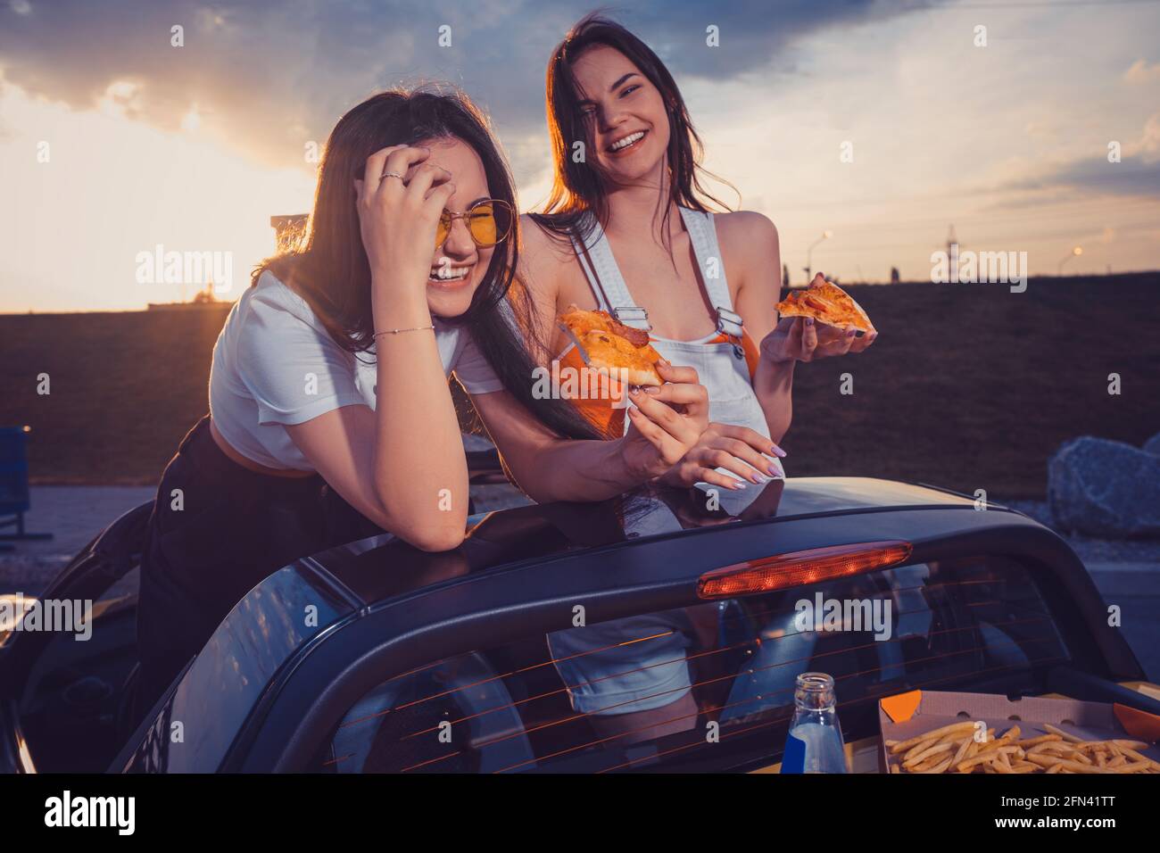 Two ladies in casual clothes eating pizza, laughing, posing in yellow car with french fries and soda in glass bottle on trunk. Fast food. Close up Stock Photo