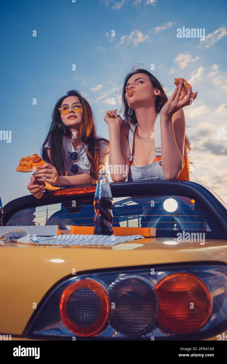 Two lovely females eating pizza while posing in yellow car cabriolet with soda in glass bottle on its trunk. Fast food. Close up, copy space Stock Photo