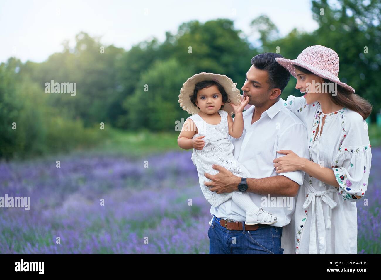 Happy family young couple posing with kid in lavender field. Little baby girl wearing straw hat sitting on hands of loving dad, looking at camera outdoors, aromatic flowers. Family, nature concept. Stock Photo