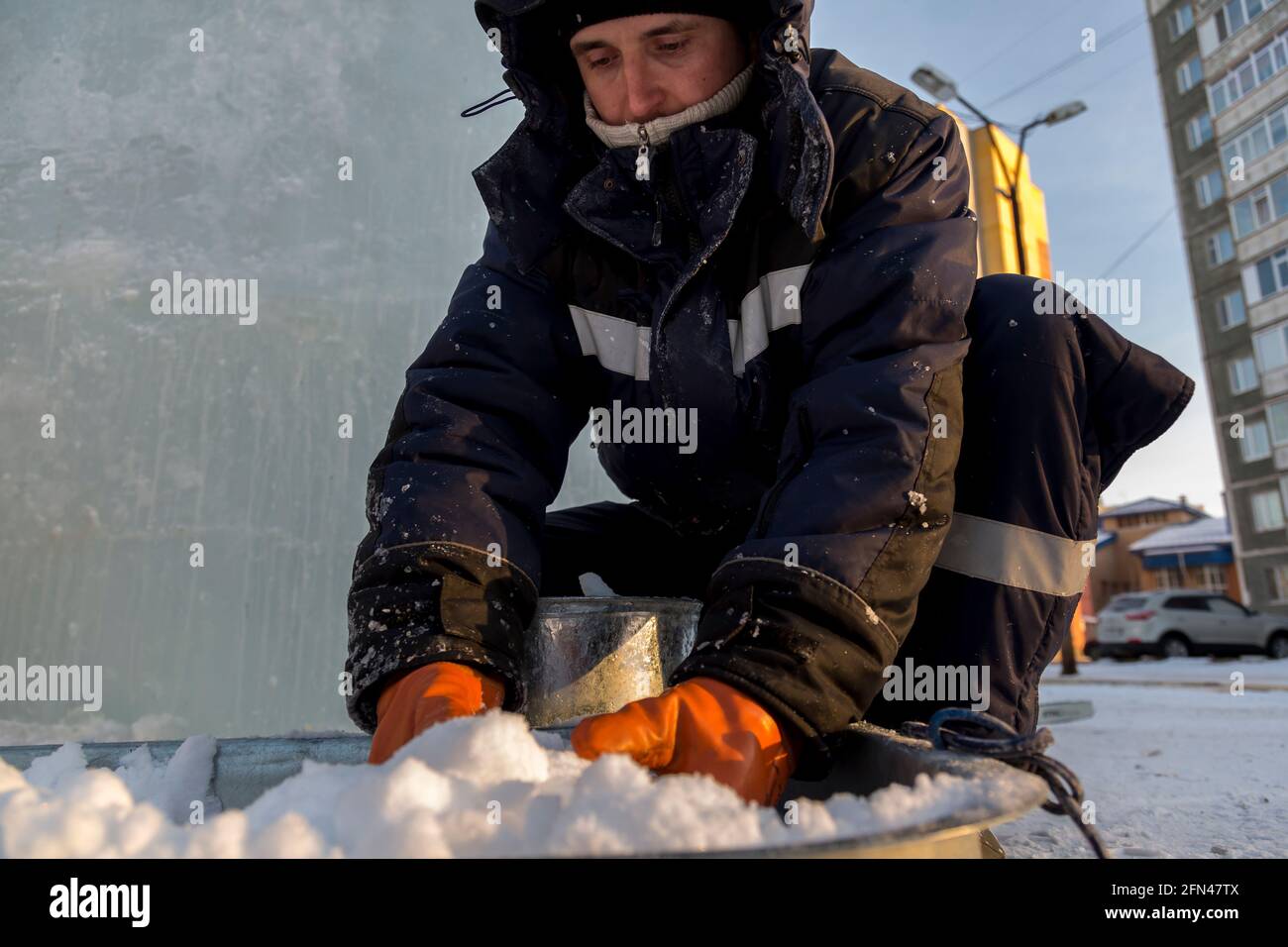 A worker assembler in winter overalls puts wet snow from a trough into a metal bucket Stock Photo
