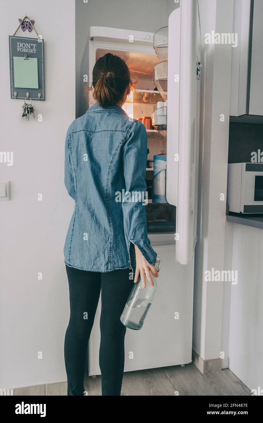 girl inside the kitchen opening the fridge door and looking inside while grabbing a bottle of water Stock Photo