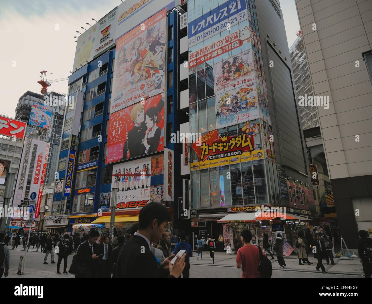 People walking in the streets of Akihabara. Businessmen and tourists stroll using smartphones in front of skyscrapers with huge manga billboards. Stock Photo