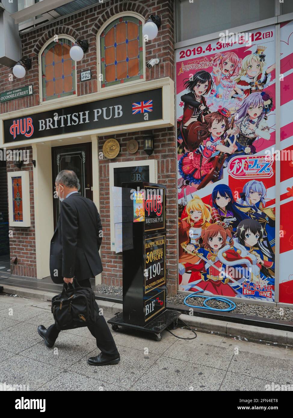 Old businessman strolls through the streets of Akihabara Electric Town. Elderly man passes in front of a fake Japanese British Pub next to a billboard Stock Photo