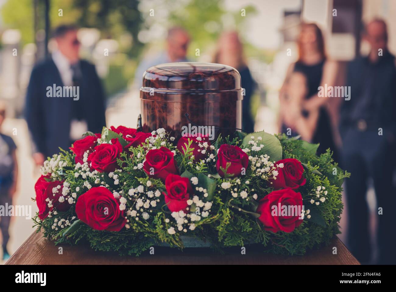 Funerary urn with ashes of dead and flowers at funeral. Burial urn decorated with flowers and people mourning in background at memorial service, sad a Stock Photo