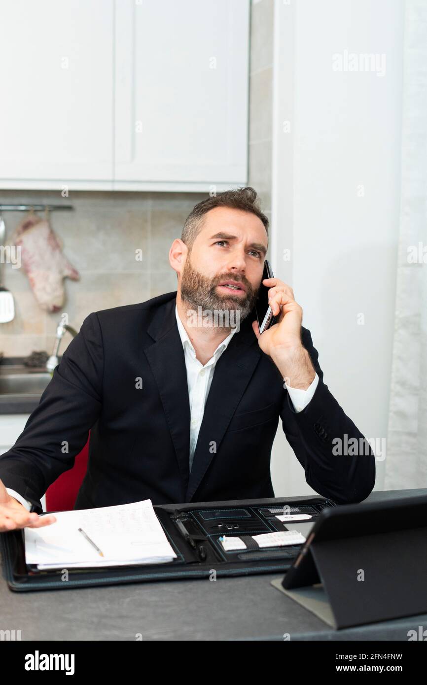 Busy young businessman calls and takes notes in the kitchen. Handsome man working at home. Smart working with tablets and sheets in covid pandemic. Stock Photo