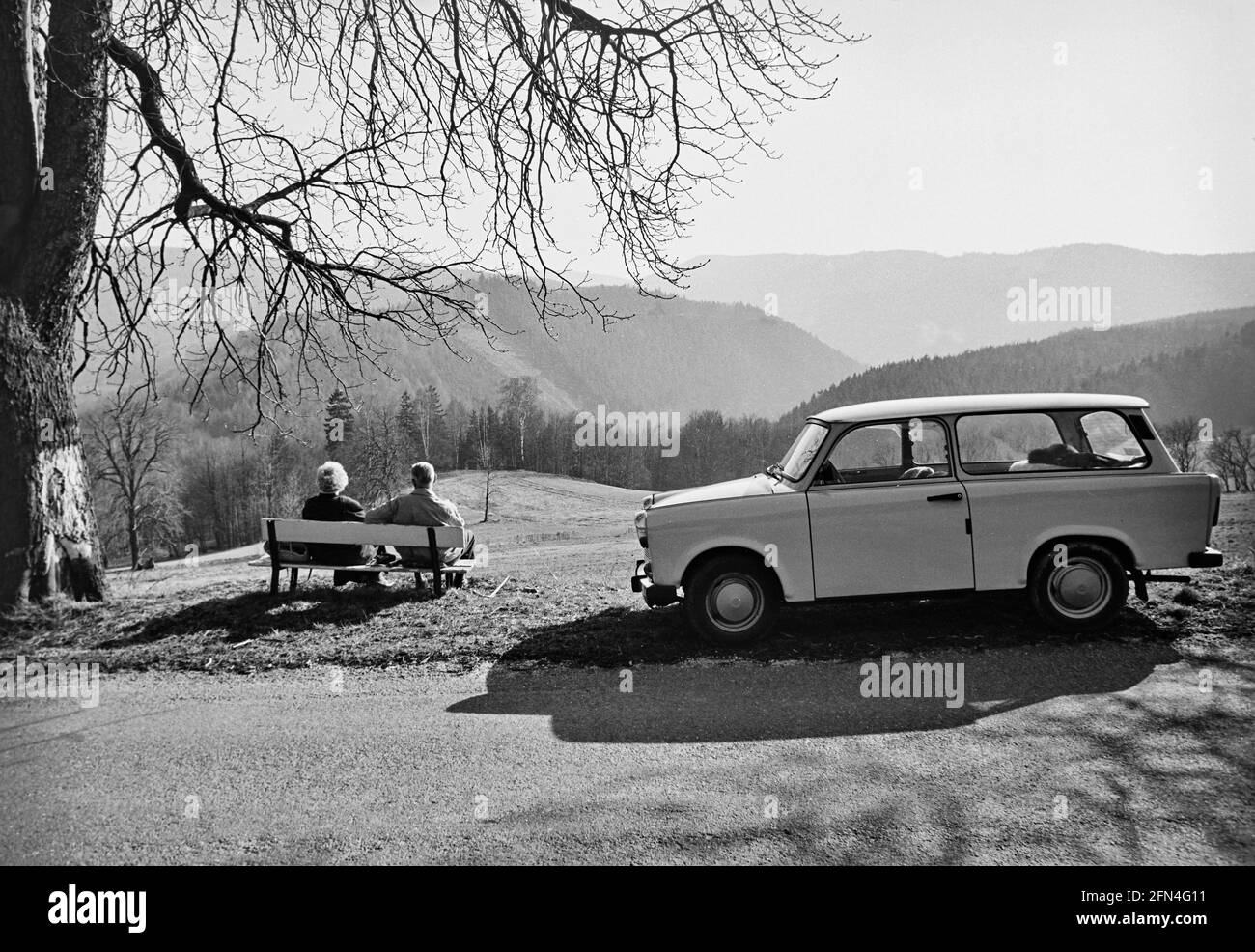 A couple enjoys the view of the wooded hills of the Thuringian Forest on a bench. The Trabant Variant stands at heel. 03.1990 - Christoph Keller Stock Photo