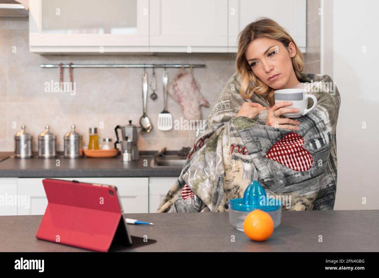 Sick blonde woman watches a tv series on the tablet. Girl fights the cold virus with a blanket over her shoulders and drinking hot herbal tea. Stock Photo