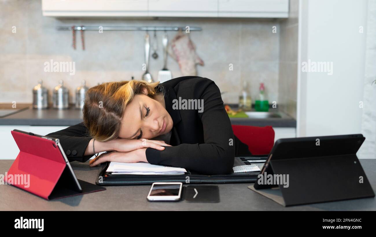 Tired business woman sleeps surrounded by tablets. Beautiful woman sleeps on the kitchen table after work from home. Smartwork during covid19 lockdown Stock Photo