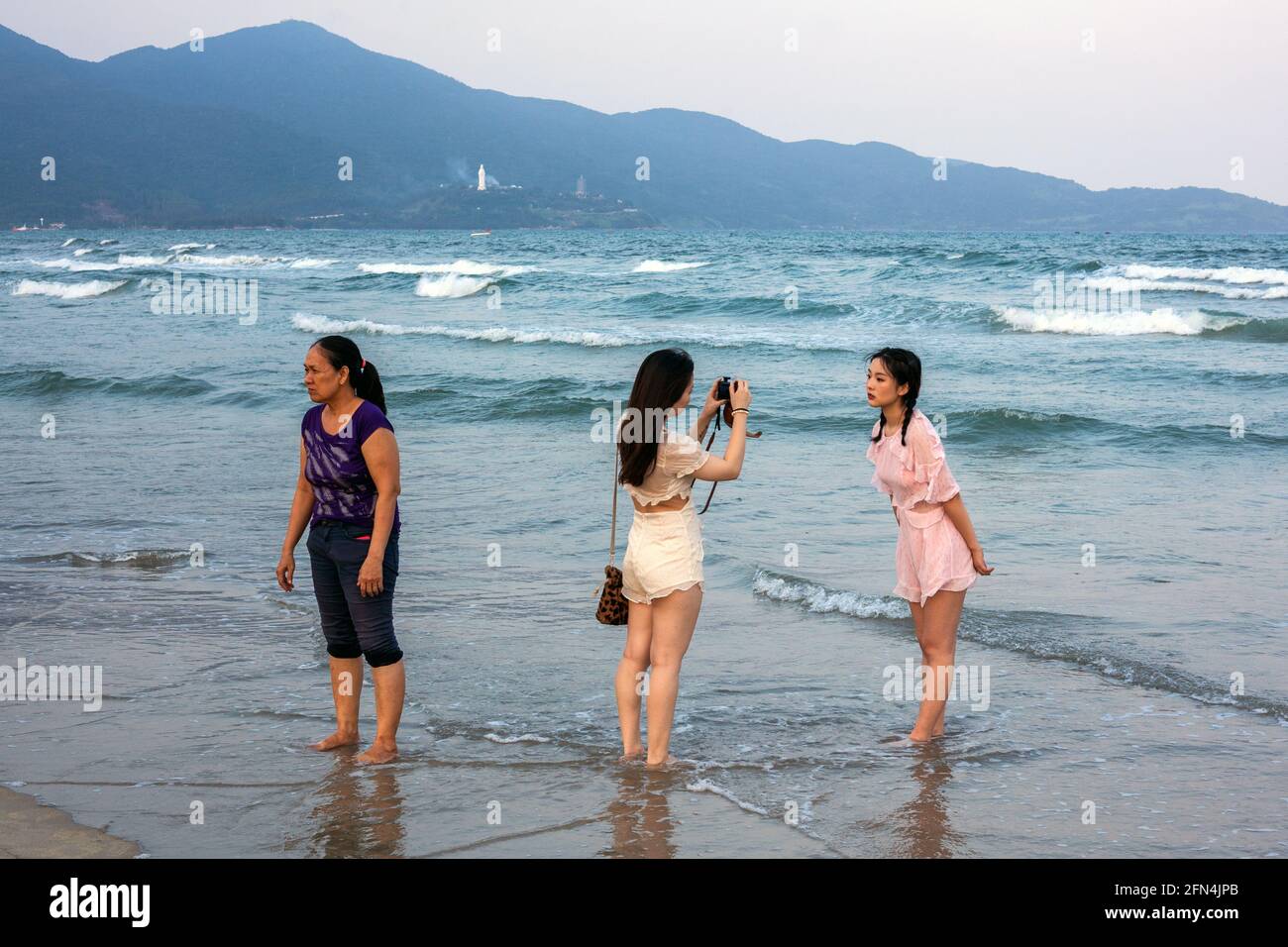 Female takes photograph of attractive friend standing in the sea at My Khe Beach, Da Nang, Vietnam Stock Photo