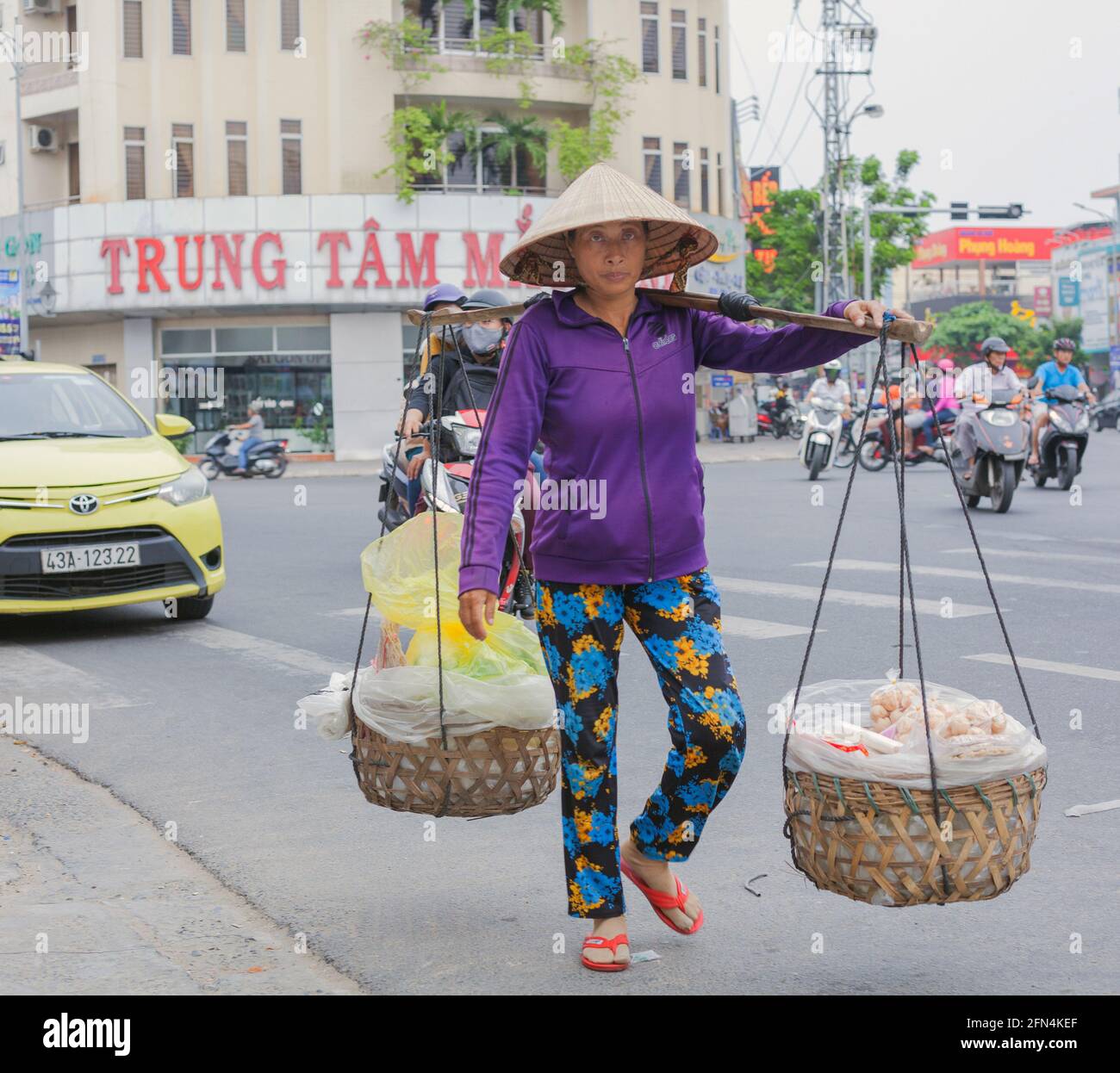 Vietnamese street vendor wearing conical hat carries food snacks in twin baskets with shoulder pole, Da Nang, Vietnam Stock Photo