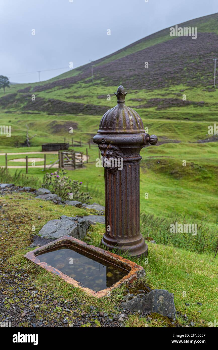 Old rusty cast iron drinking fountain with decorative Lion Head pattern and a ceramic basin sunk into ground at Wanlockhead in the Leadhills Stock Photo