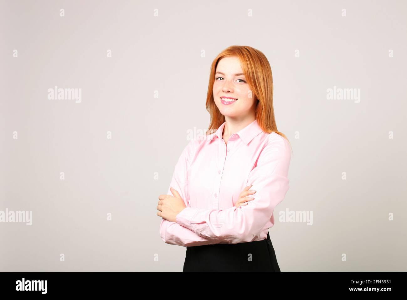Beautiful businesswoman wearing formal wear, black skirt & pink blouse standing with arms folded on chest. Attractive woman w/ red hair & light make u Stock Photo