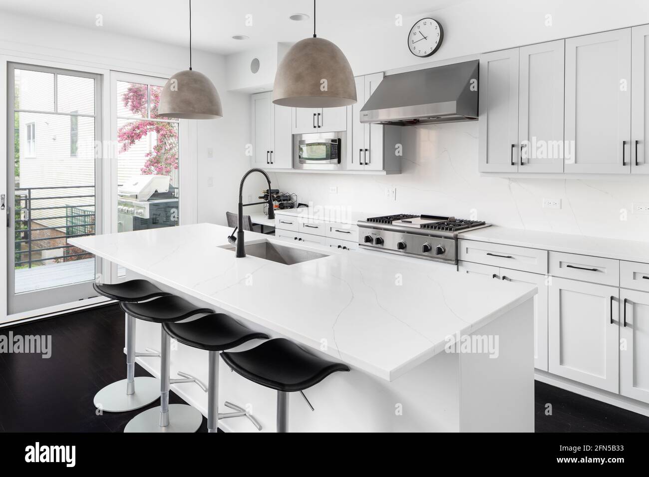 An elegant white kitchen with Thermador stainless steel appliances, large island with a granite countertop, and modern lights hanging above. Stock Photo