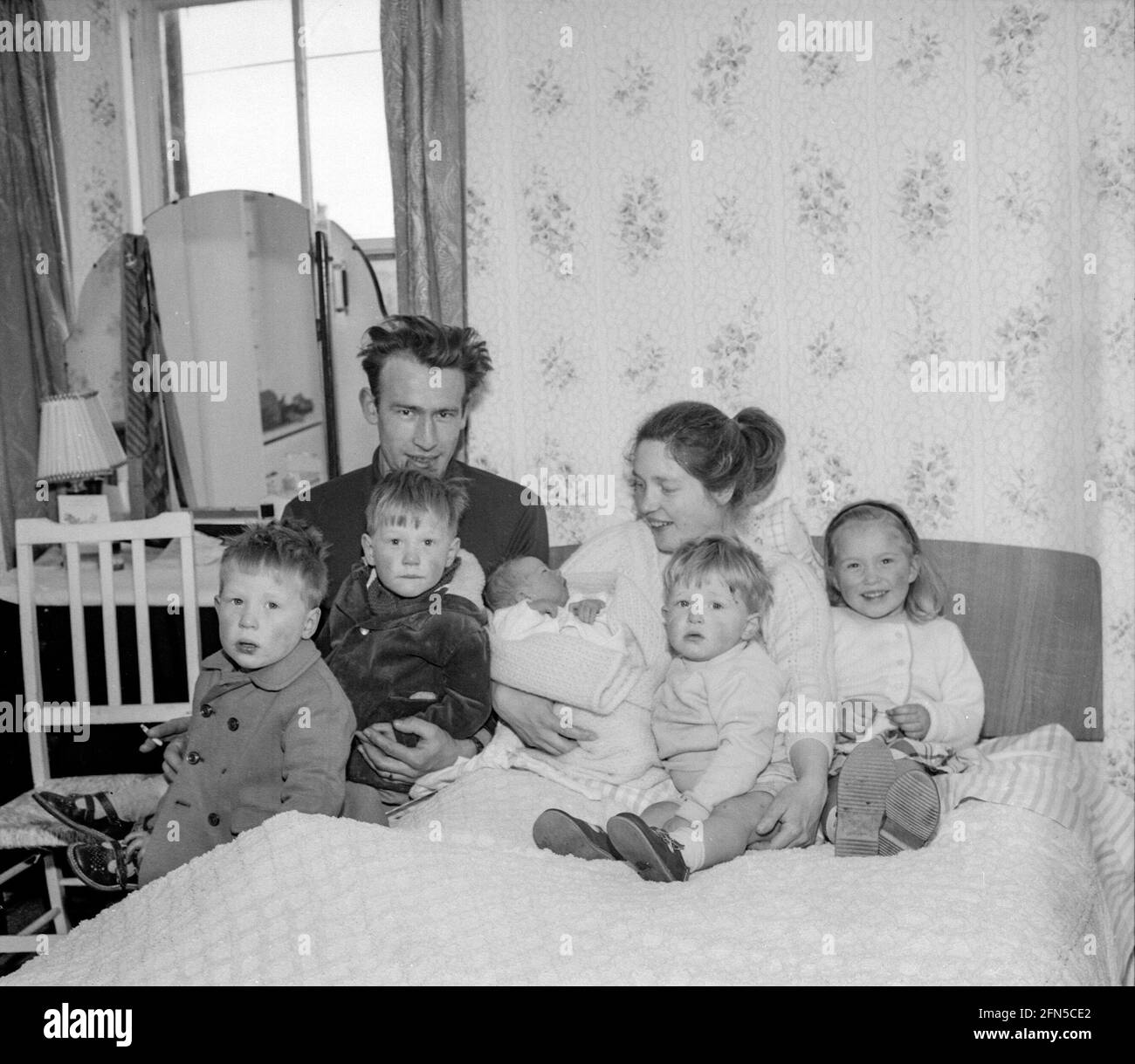 A young couple and their five young children sit on a bed in the room they all share typifies the British housing crisis in the 1950s. In the decades after World War II a huge programme of Council house building took place to remedy this crisis. Stock Photo