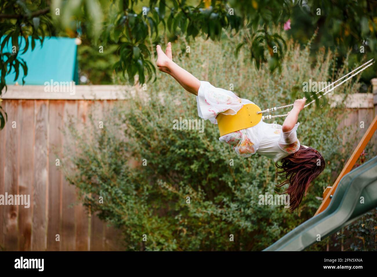 a barefoot child swings high on a playlet in a backyard garden Stock Photo