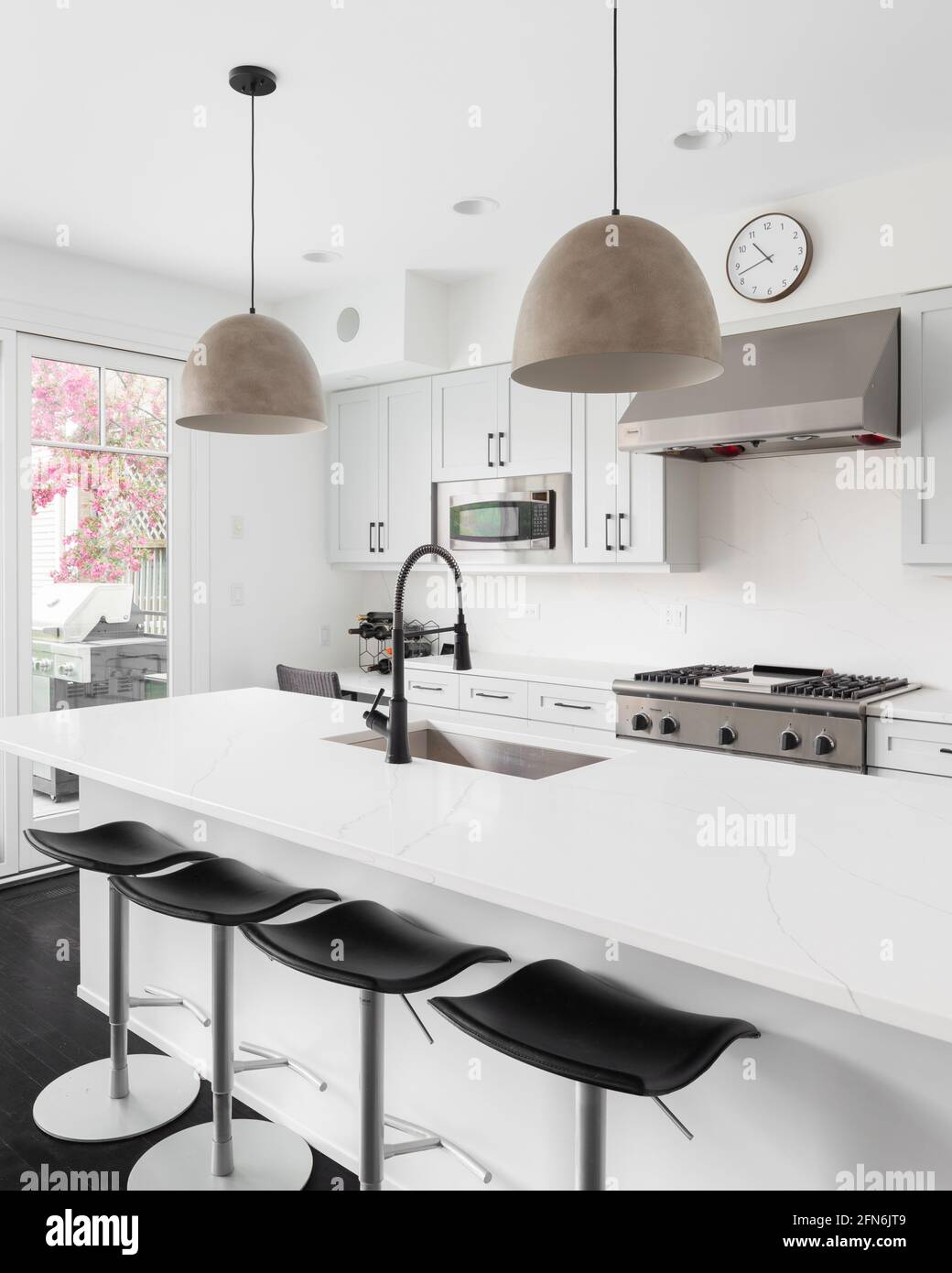 An elegant white kitchen with Thermador stainless steel appliances, large island with a granite countertop, and modern lights hanging above. Stock Photo