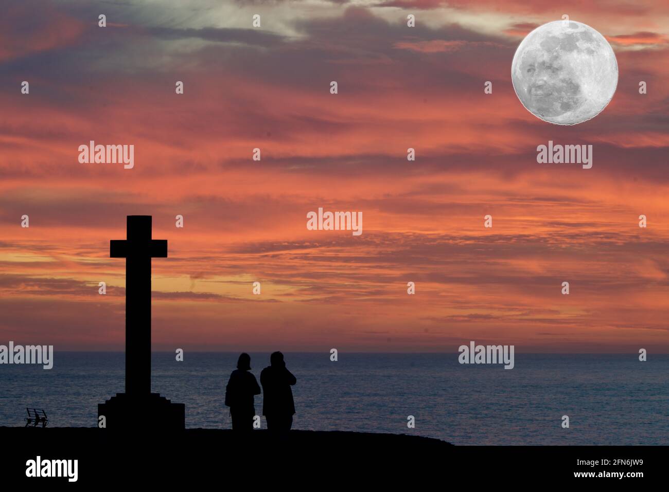 The man in the moon is watching a couple beside a cross in a red sundown Stock Photo