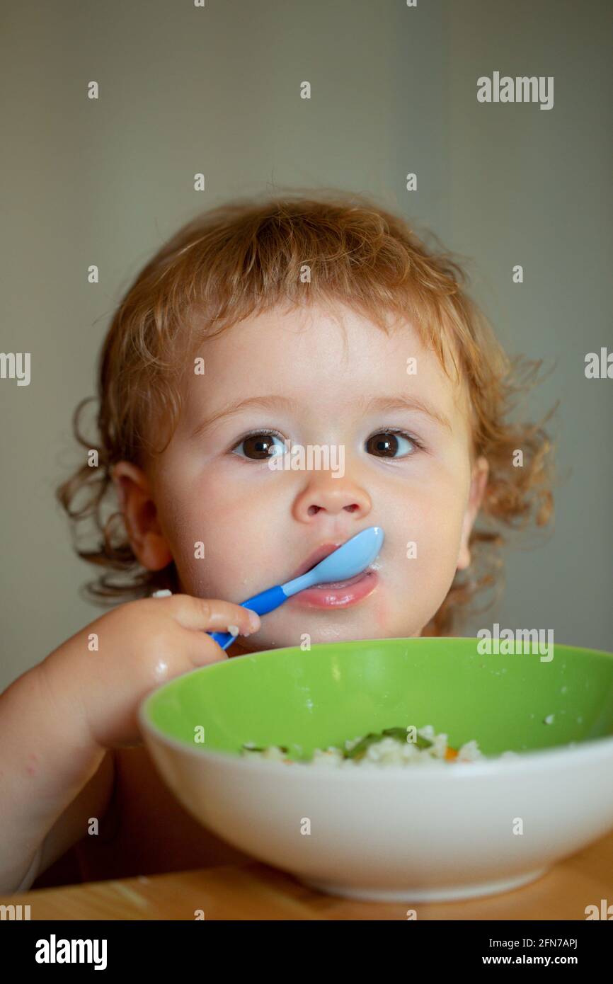 Funny baby eating food himself with a spoon on kitchen. Child nutrition concept. Funny child face closeup. Stock Photo