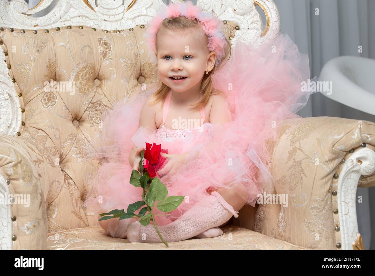 A little girl in an elegant pink dress with a red rose sits in a chair ...
