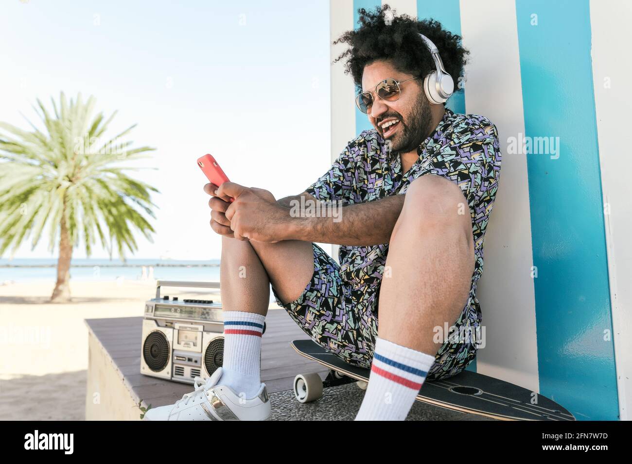 Afro Latin man having fun with mobile smartphone and listening music with headphones and vintage boombox stereo on tropical beach during vacation time Stock Photo