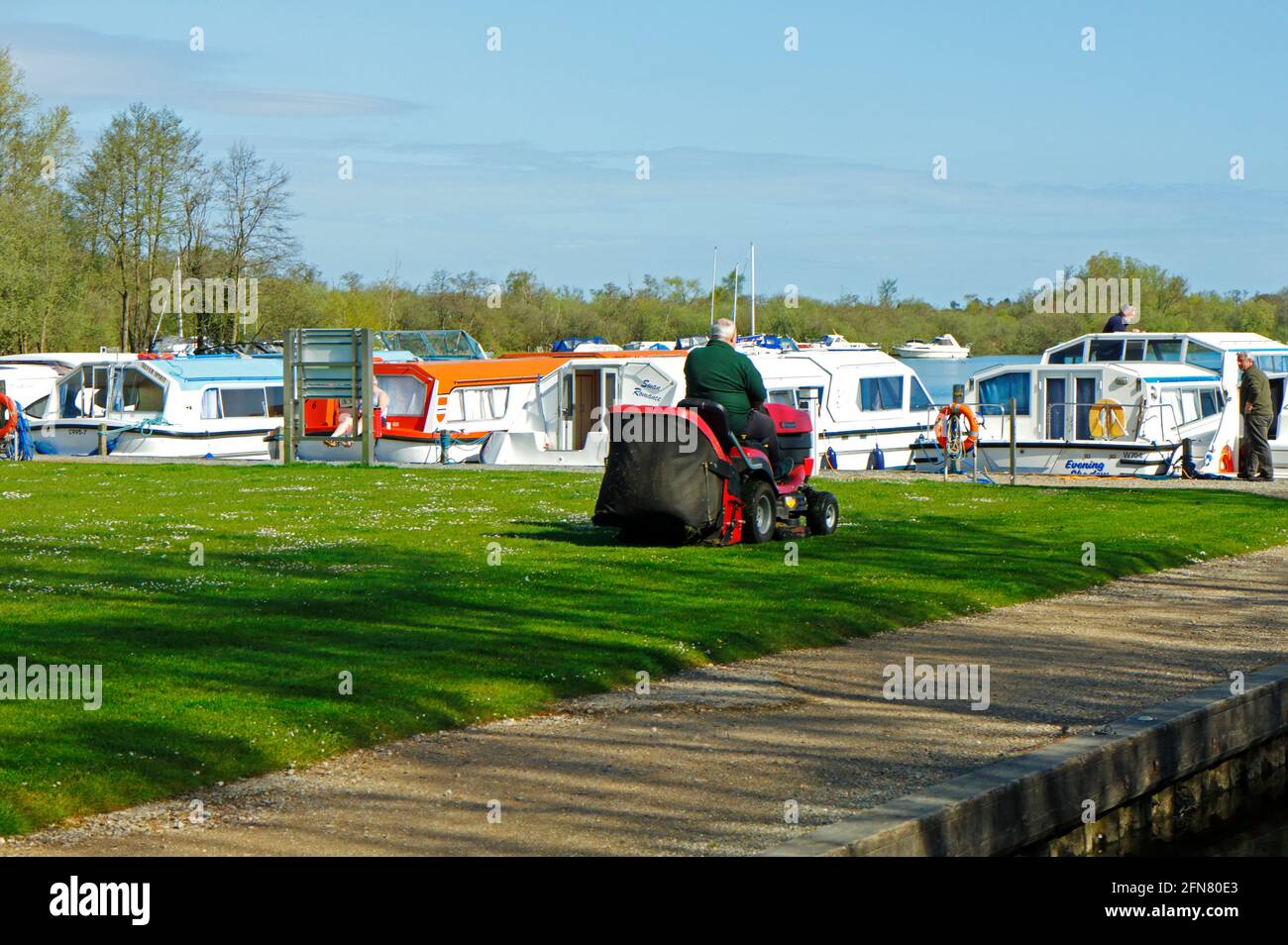 A motorized sit-on grass cutter with operator on the green by Malthouse Broad at Ranworth, Norfolk, England, United Kingdom. Stock Photo