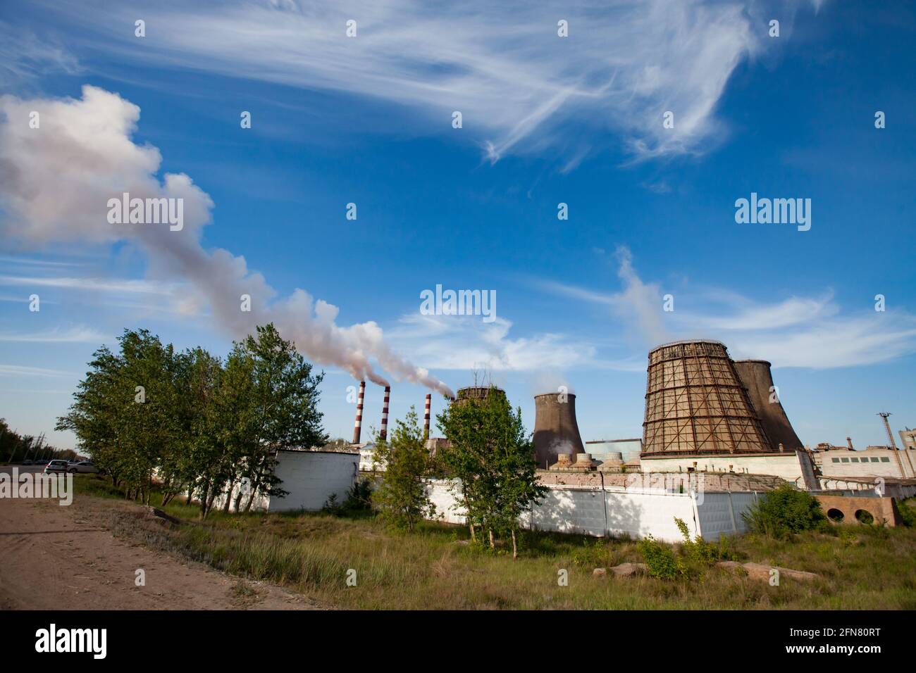 Pavlodar thermal electric station. Cooling towers and smoke stack with white smoke. Green grass, trees, blue sky with clouds. Stock Photo