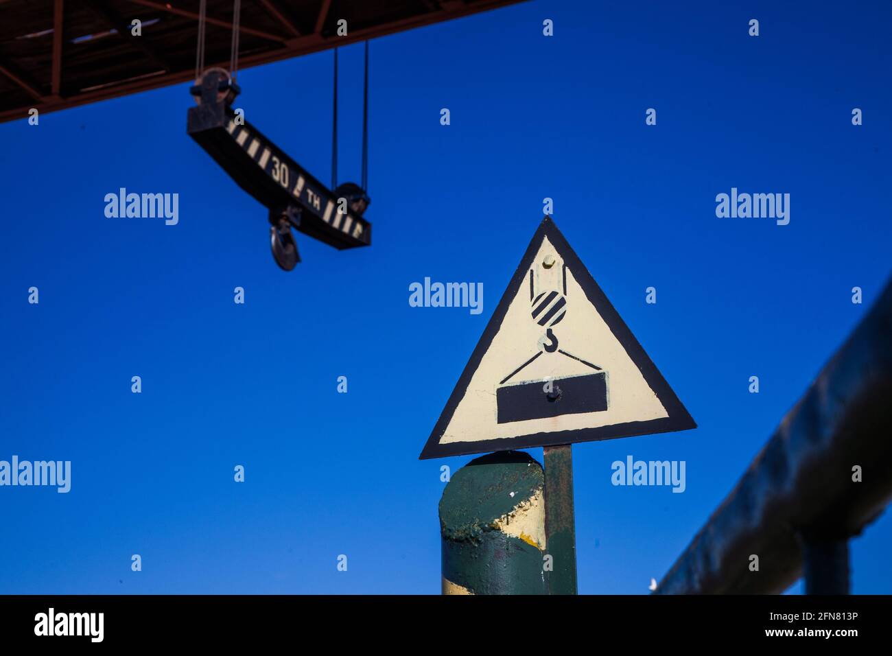 Self-made vintage warning road sign 'Crane working' against deep blue sky. Overhead crane on background, blurred. Stock Photo