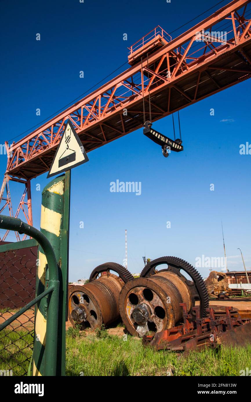 Overhead crane and giant gears of walking excavator's winch on the ground and grass. Vintage warning road sign 'Crane working' against deep blue sky. Stock Photo