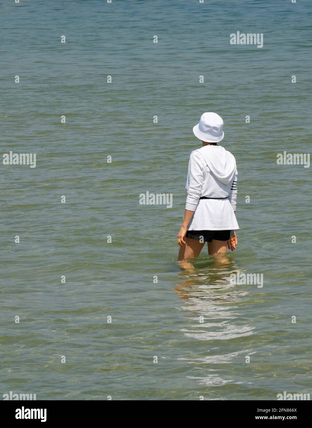 A young woman dressed in a white top and hat, standing in shallow sea water on a sunny day Stock Photo