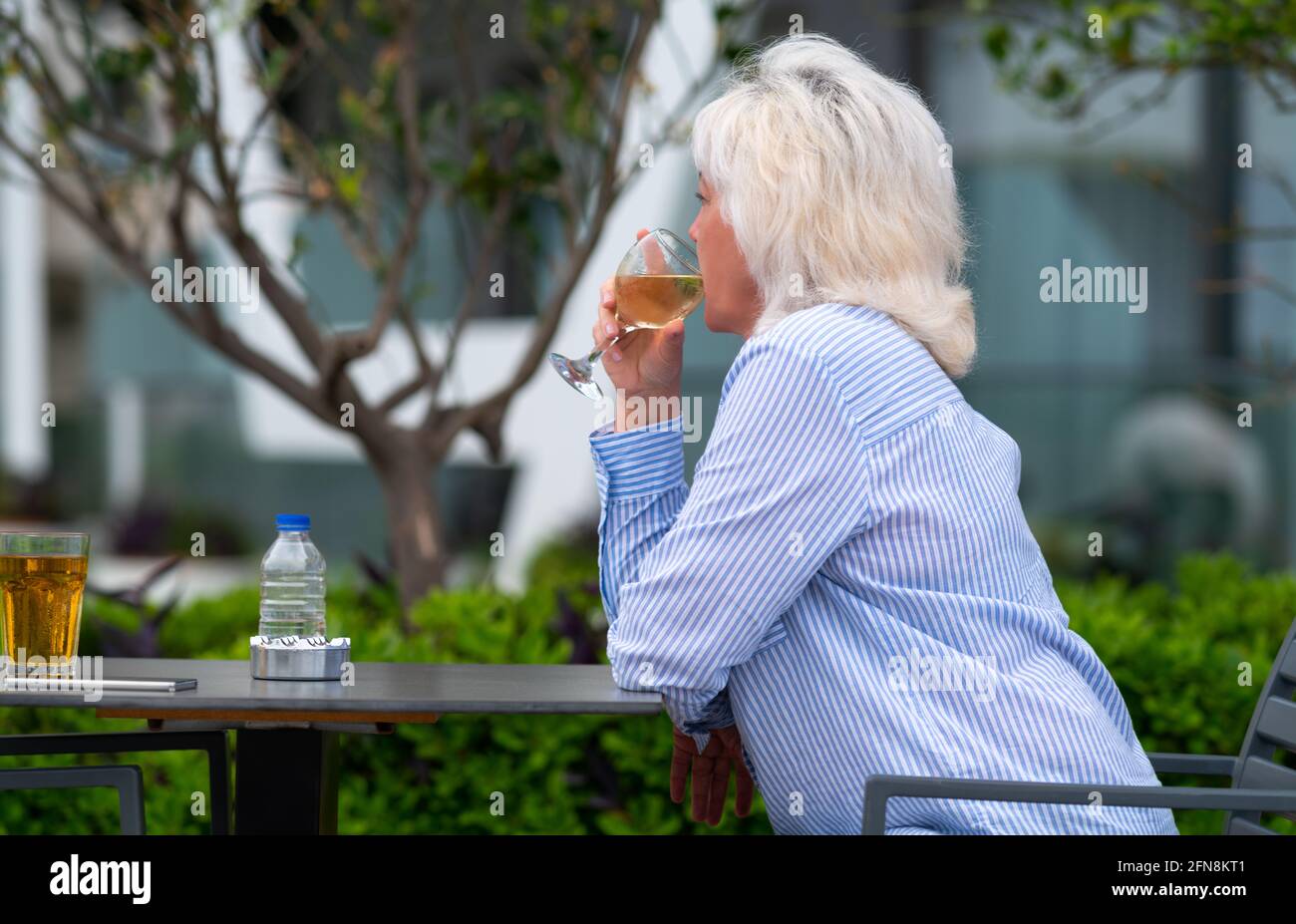 Woman sitting quietly sipping a glass of white wine outdoors at a table as she enjoys a relaxing day in a close up low angle upper body side view with Stock Photo