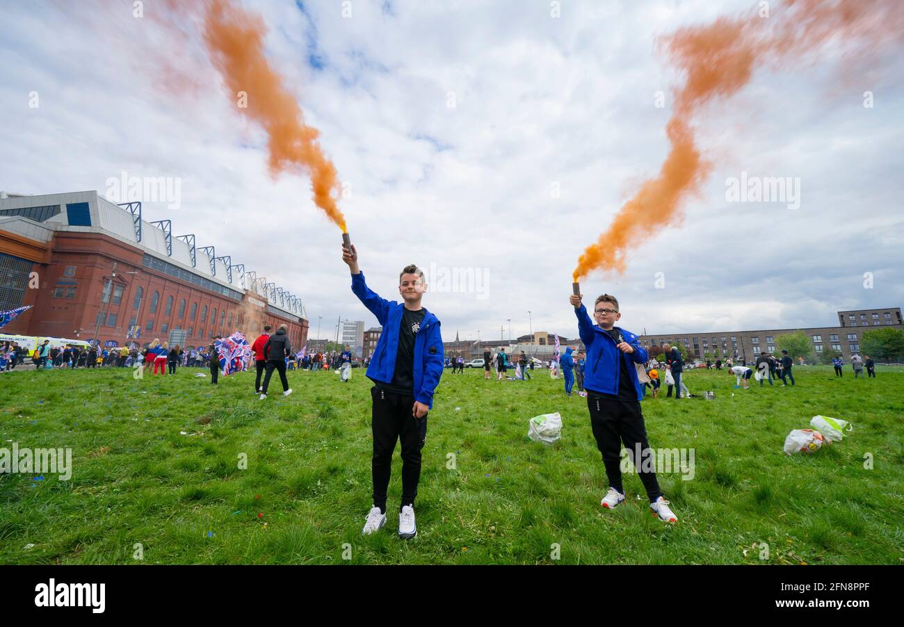 Glasgow, Scotland, UK. 15 May 2021. Thousands of supporters and fans of Rangers football club descend on Ibrox Park in Glasgow to celebrate winning the Scottish Premiership championship for the 55nd time and the first time for 10 years. Smoke bombs and fireworks are being let off by fans tightly controlled by police away from the stadium entrances.Pic; Young fans with orange smoke flares. Iain Masterton/Alamy Live News Stock Photo