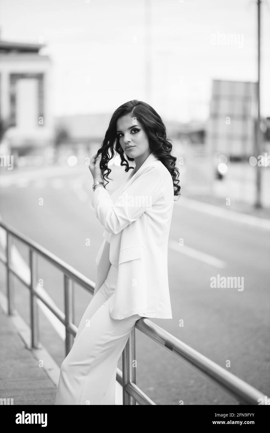 Grayscale of a gorgeous Caucasian female in an elegant outfit leaning on an urban street fence Stock Photo