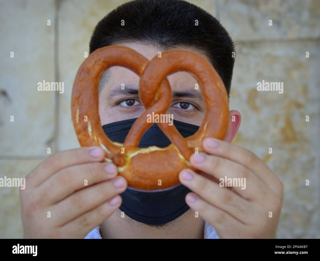 Young Caucasian baker with black face mask holds a traditional German Bavarian lye pretzel with both hands and looks through the pretzel loop. Stock Photo