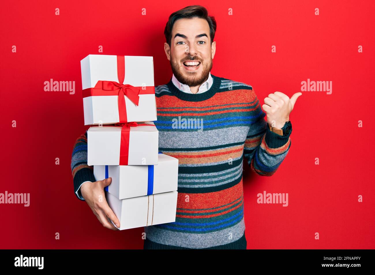 Handsome caucasian man with beard holding gifts pointing thumb up to ...