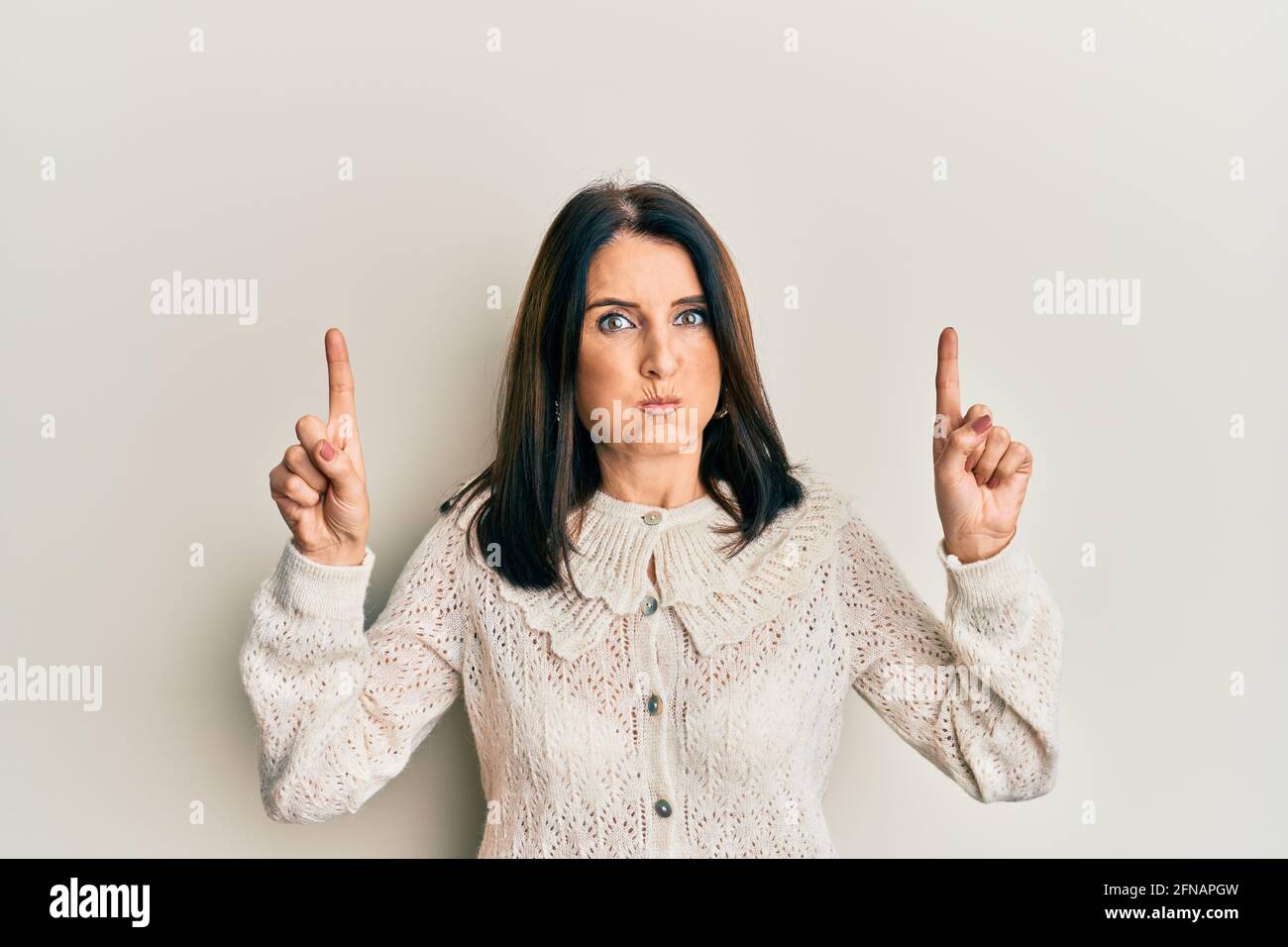 Middle age brunette woman pointing up with fingers puffing cheeks with funny face. mouth inflated with air, catching air. Stock Photo