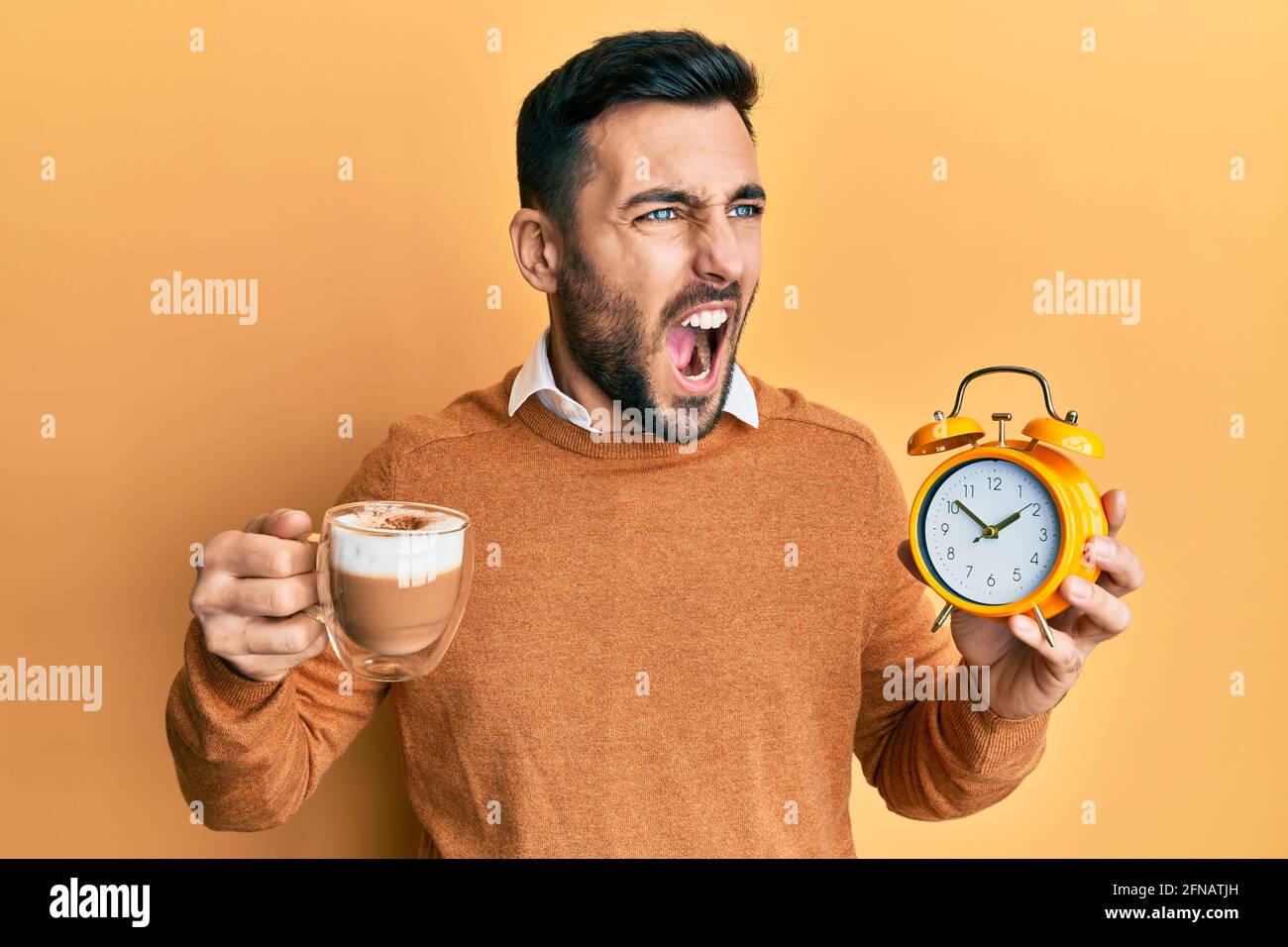 Young hispanic man holding coffee and alarm clock angry and mad