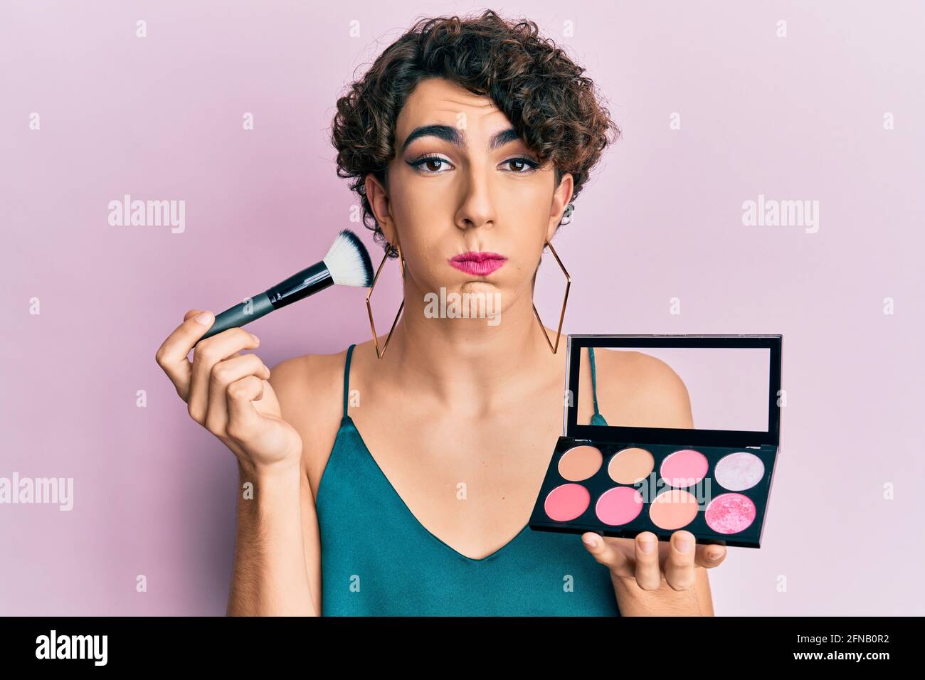 Young man wearing woman make up holding makeup brush and blush puffing cheeks with funny face. mouth inflated with air, catching air. Stock Photo