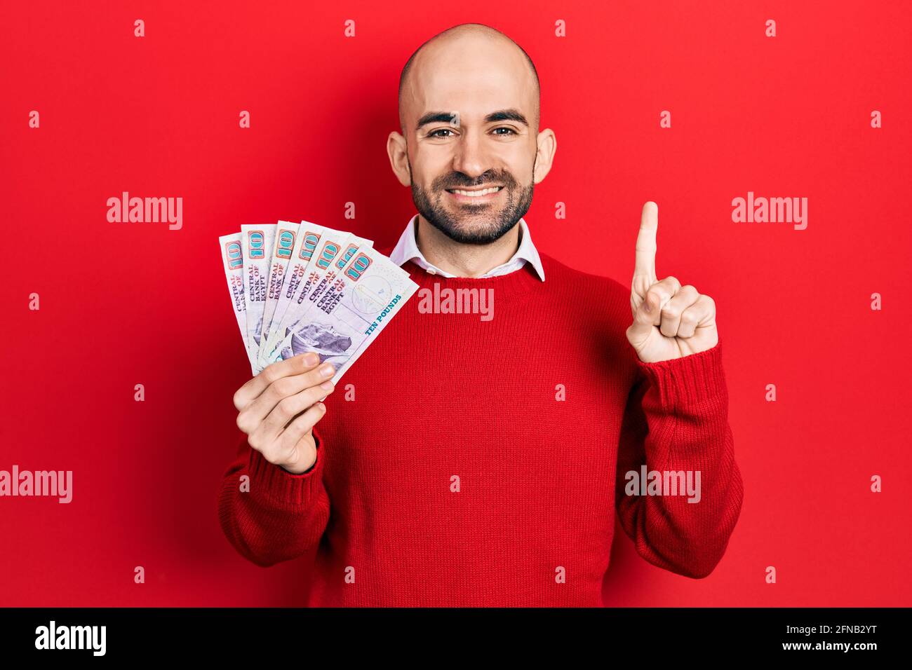 Young bald man holding egyptian pounds banknotes smiling with an idea or question pointing finger with happy face, number one Stock Photo